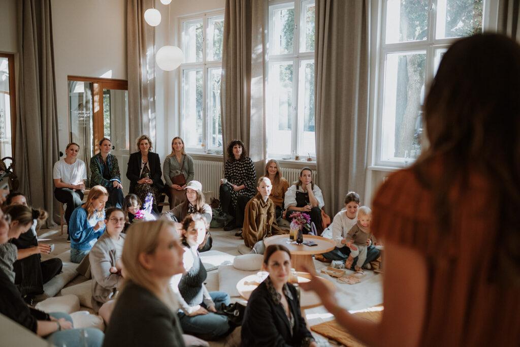 Man sieht Frauen auf dem Boden sitzend, auf Stühlen und stehend-Sie befinden sich in einem hellen Raum mit großen Fenstern und hören einem Vortrag zu.