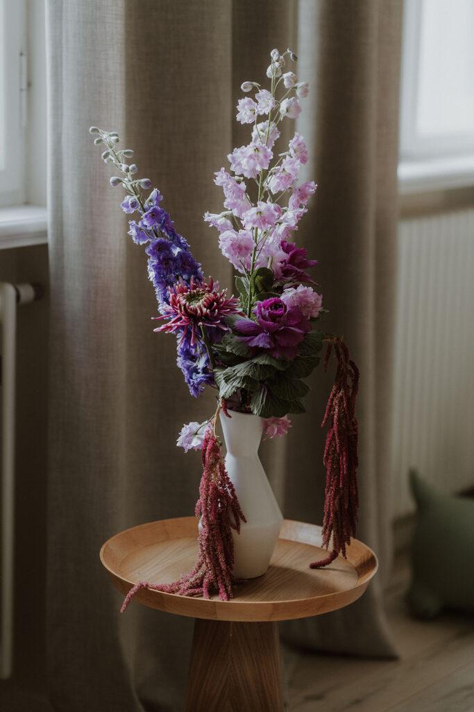 Ein großer Blumenstrauß in einer hohen Vase mit verschiedenen lilafarbenen Blumen auf einem kleine Beistelltisch aus Holz.