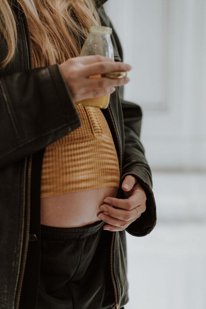 Eine Nahaufnahme einer Schwangeren, die sich an den Bauch fasst. Sie fasst sich mit einer Hand an den Bauch, die andere hält eine Glasflasche mit Orangesaft. Sie trägt ein gelbes Strickoberteil, eine Lederjacke und eine dunkle Hose.